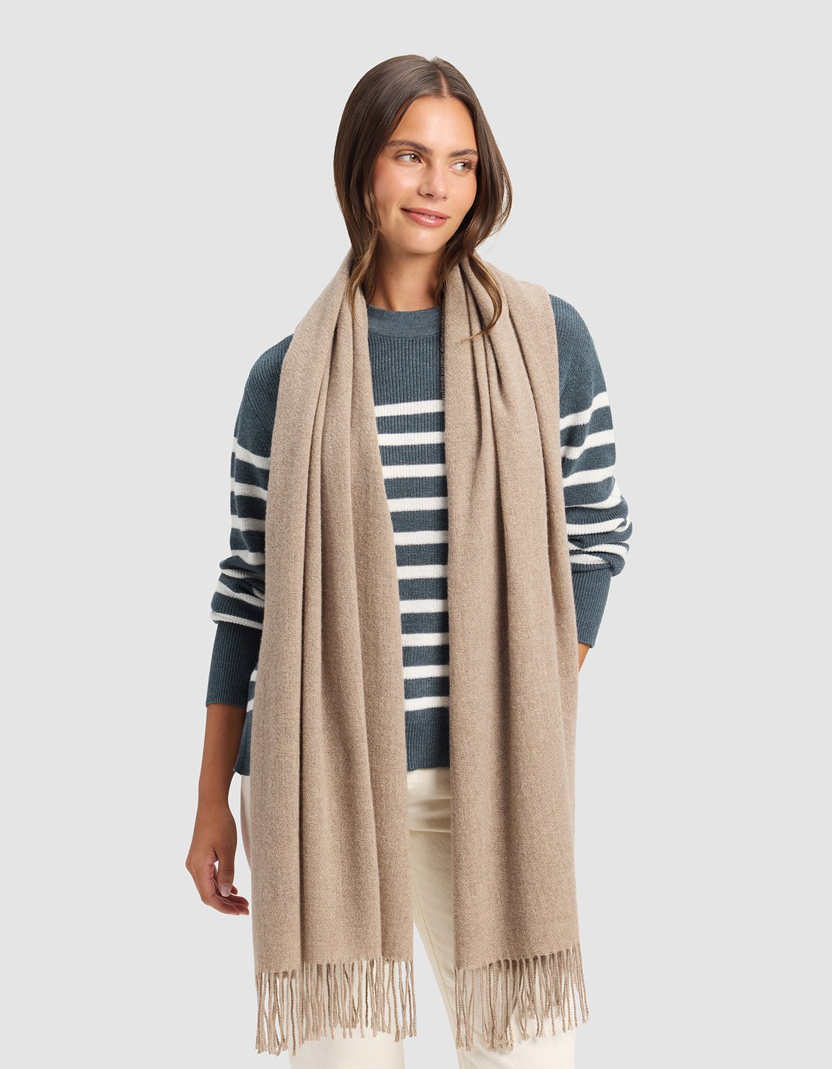 A woman stands against a light gray background wearing the Cozy Earth Amelia Wrap, a long beige scarf with fringed ends, paired with a blue and white striped sweater and cream-colored pants. |Color:Taupe