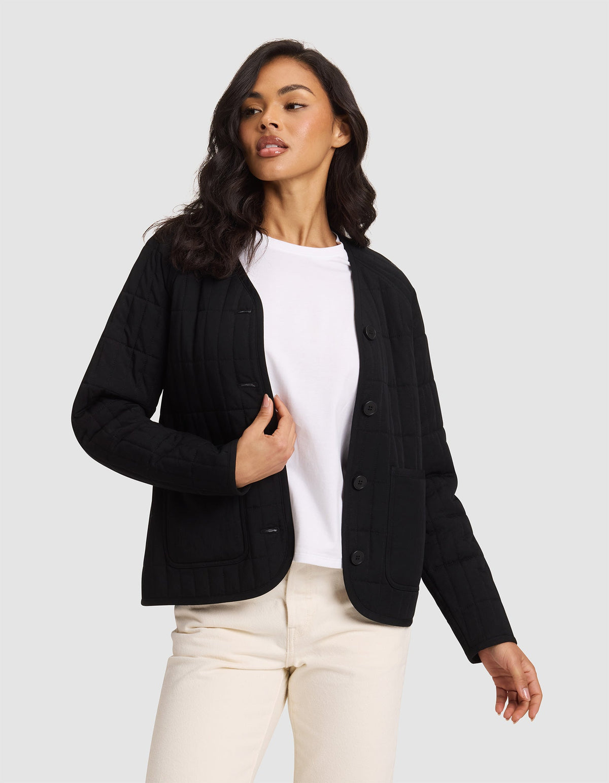 A woman with long, wavy dark hair stands against a light gray background wearing the Cozy Earth Women's Andie Quilted Jacket in black over a white t-shirt and cream-colored pants, looking slightly to the side.|Color:Jet Black
