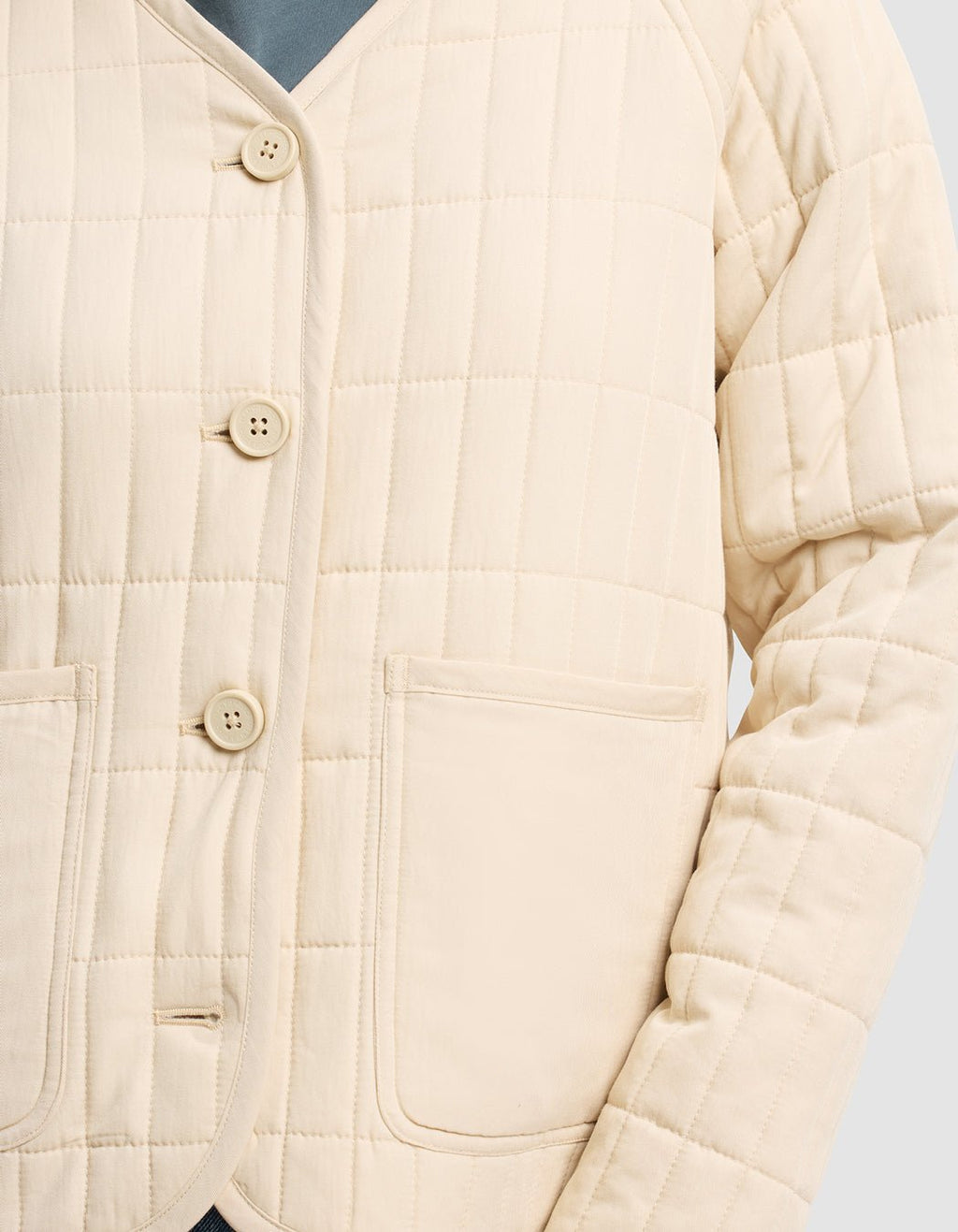 A close-up of the Women's Andie Quilted Jacket by Cozy Earth, shown in cream with grid pattern stitching, four buttons, and two front pockets, worn over a blue top.|Color:Sesame