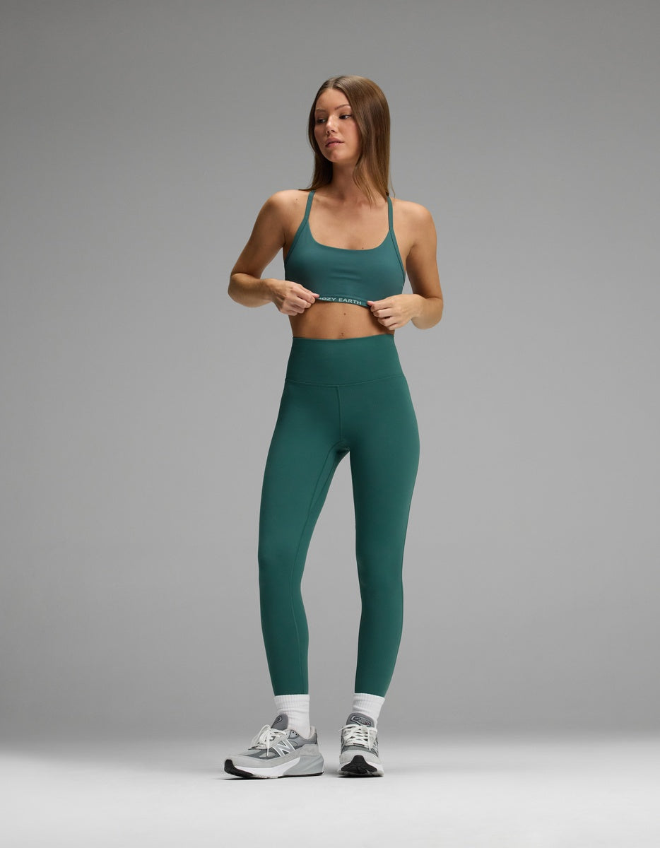 A woman wearing a dark green Cozy Earth Women’s Balance Bra and matching high-waisted leggings, white crew socks, and gray athletic shoes.|Color:Uniform