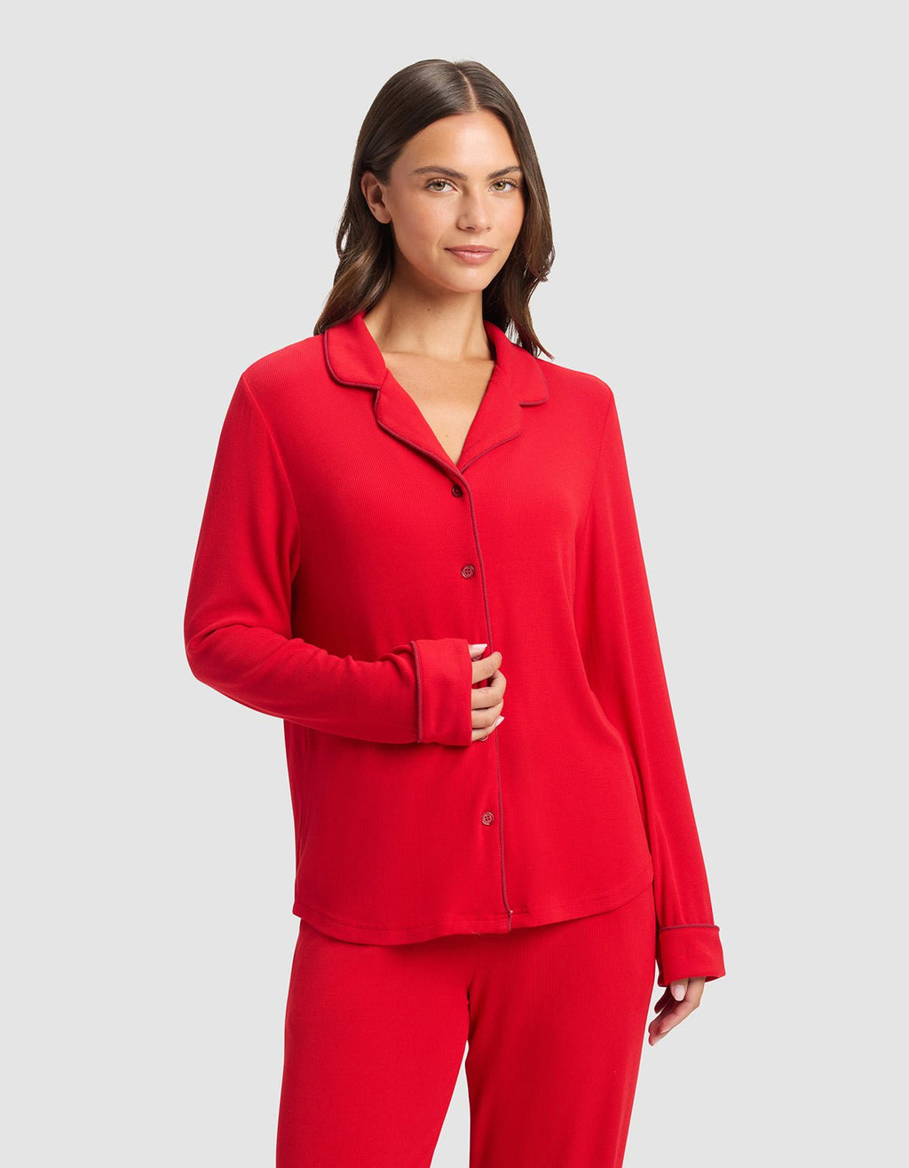 A woman with long brown hair stands against a light gray background, wearing the Cozy Earth Women's Bamboo Rib-Knit Classic Long Sleeve Pajama Top in red and matching pants. She smiles gently with one hand on her abdomen.|Color:Crimson