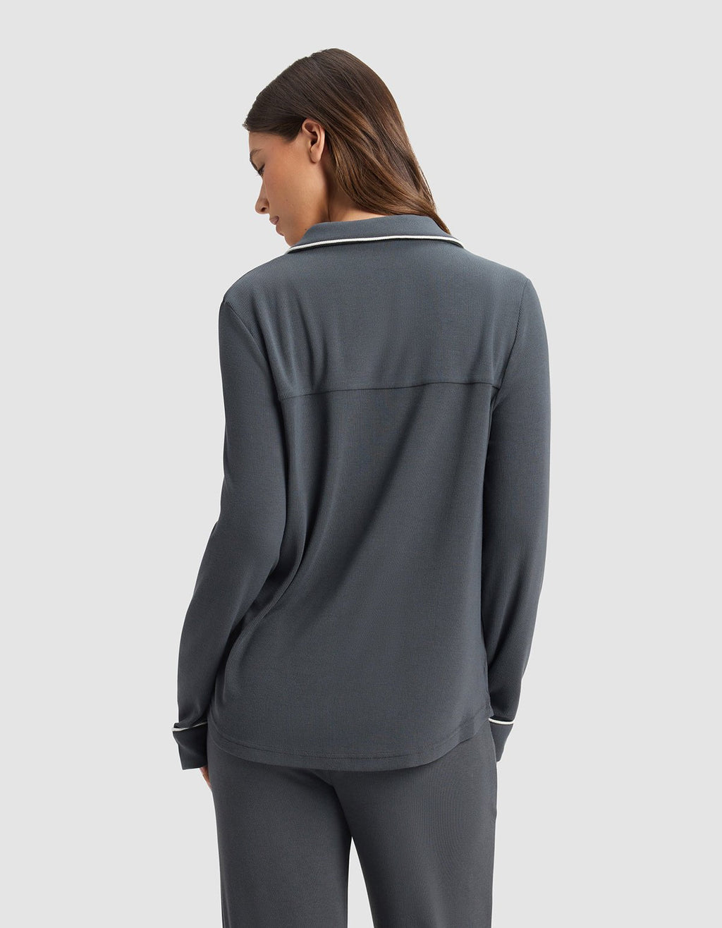 A woman with long brown hair stands facing away, wearing the Cozy Earth Women's Bamboo Rib-Knit Classic Long Sleeve Pajama Top in dark gray against a light gray background.|Color:Graphite