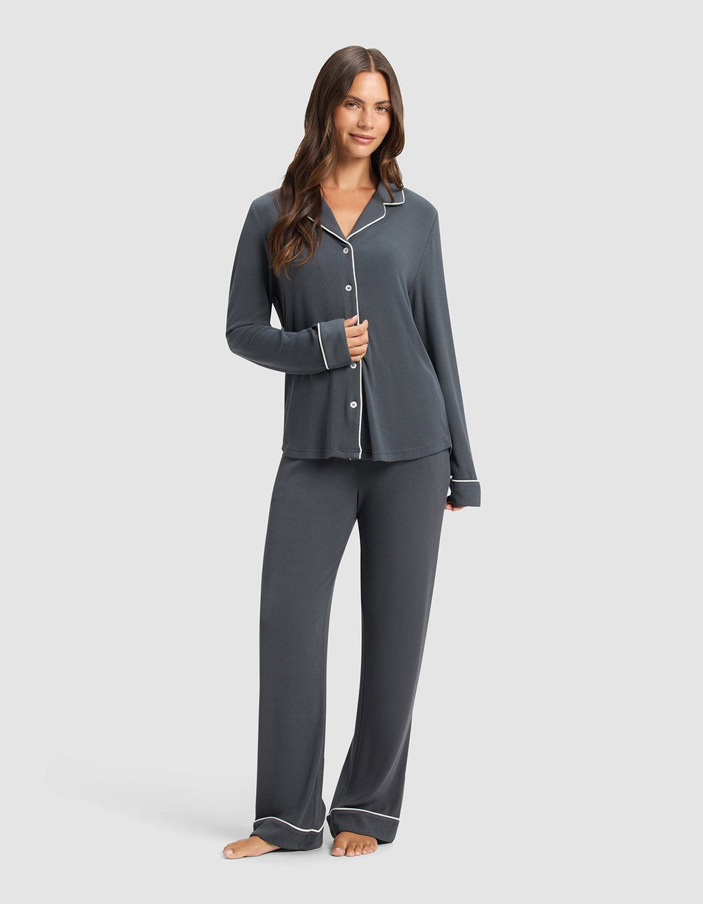 A woman with long, wavy hair and a relaxed expression stands barefoot, wearing Cozy Earth's Women's Bamboo Rib-Knit Classic Pajama Pant in dark gray with white piping, paired with a matching long-sleeve button-up top.|Color:Graphite