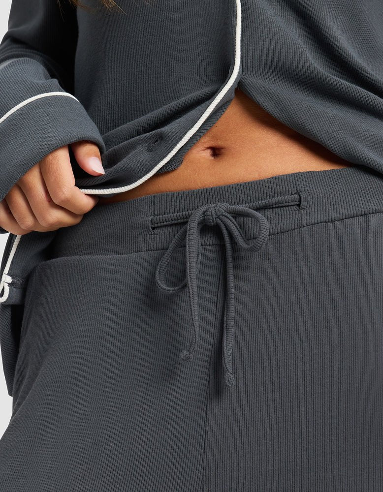 Close-up of a person wearing Cozy Earth's Women's Bamboo Rib-Knit Classic Pajama Pant in dark gray with a drawstring waist and matching long-sleeve top, slightly lifting the top to reveal part of their midriff.|Color:Graphite