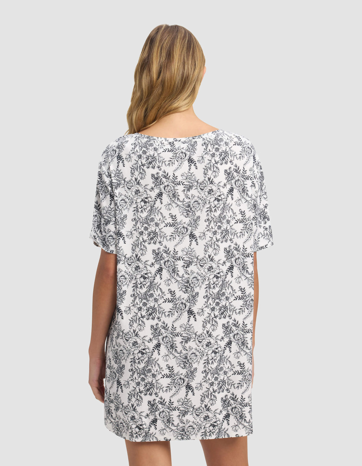 A woman with blonde hair stands facing away, wearing the Cozy Earth Women's Bamboo Stretch-Knit Sleep Dress in white with a black floral pattern on a light gray background.|Color:Jet Black Toile