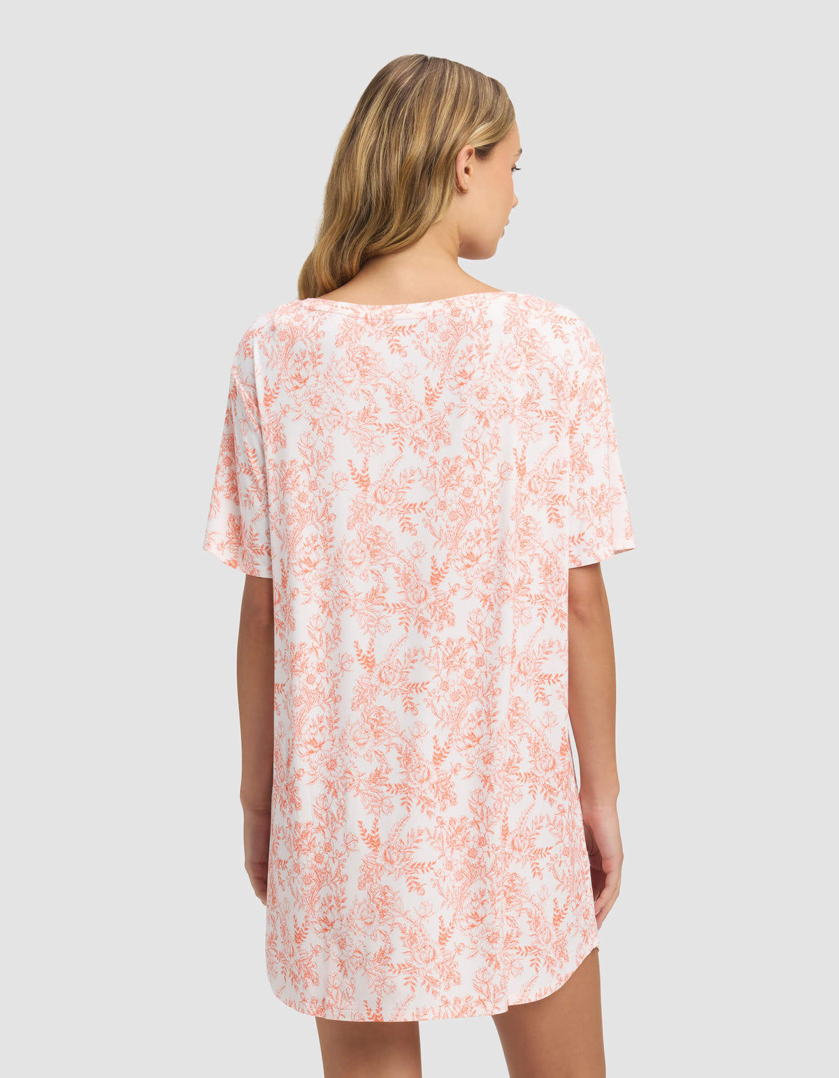 A woman with long blonde hair is seen from the back wearing the Cozy Earth Women’s Bamboo Stretch-Knit Sleep Dress, a loose short-sleeved outfit in a light pink and white floral pattern, set against a plain gray background.|Color:Melon Toile
