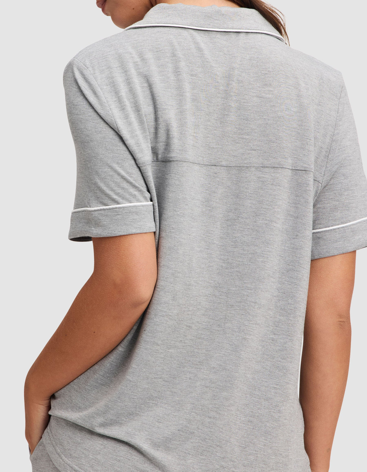 A woman is shown from the back wearing the Cozy Earth Women's Bamboo Stretch-Knit Short Sleeve Pajama Set in light gray, featuring a collared top with white trim and short sleeves, with her left arm bent and hand in her pocket. |Color:Grey