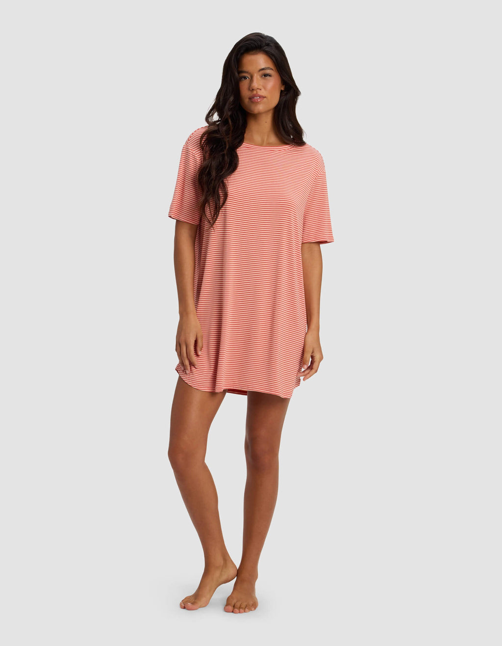 A woman stands barefoot in a Cozy Earth Women's Bamboo Stretch Knit Sleep Dress, featuring loose-fitting short sleeves and pink and white stripes, set against a plain light gray background.|Color:Sunset Mini Stripe