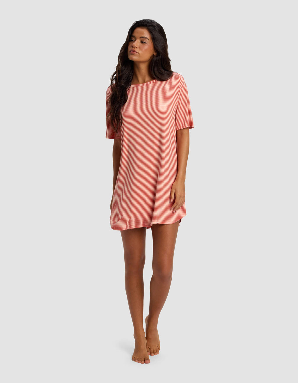 A woman with long dark hair stands barefoot against a light gray background, wearing the Cozy Earth Women's Bamboo Stretch Knit Sleep Dress—a loose-fitting, short-sleeved style in light pink and white stripes.|Color:Sunset Mini Stripe