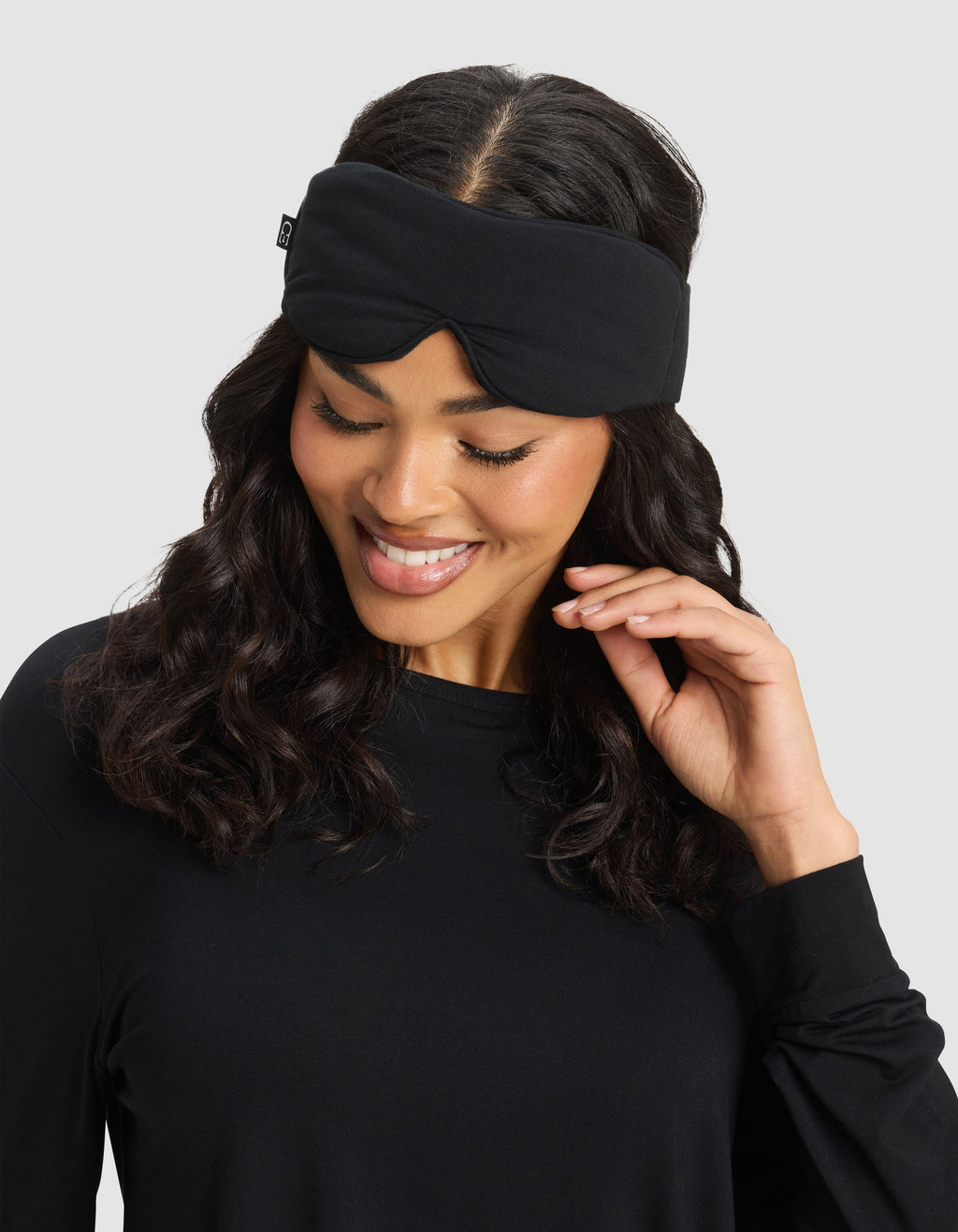 A woman with wavy dark hair smiles while wearing the Cozy Earth Bamboo Stretch-Knit Sleep Mask pushed up on her forehead and a black long-sleeve top, touching her hair against a plain light gray background.|Color:Black
