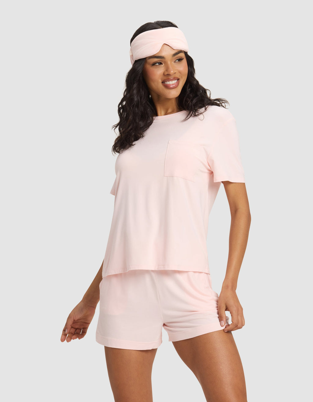 A woman stands smiling against a light gray background wearing a light pink pajama set with shorts and a Cozy Earth Bamboo Stretch-Knit Sleep Mask in matching pink on her head.|Color:Blossom
