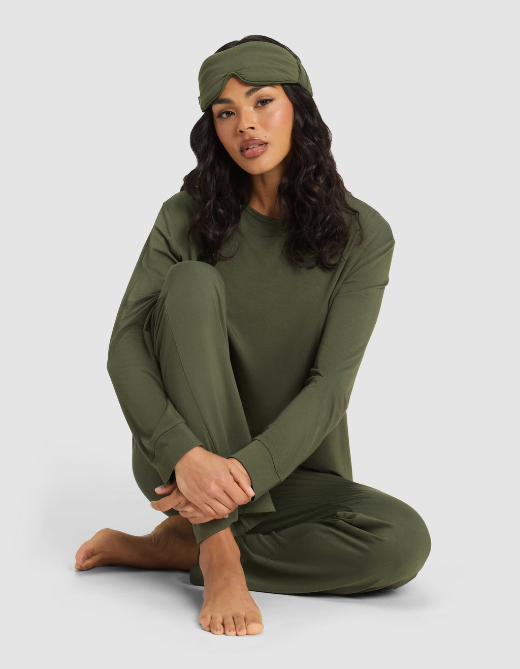 A woman with long dark hair sits barefoot on the floor, relaxed against a plain light background, wearing an olive green pajama set and the Cozy Earth Bamboo Stretch-Knit Sleep Mask on her head.|Color:Olive