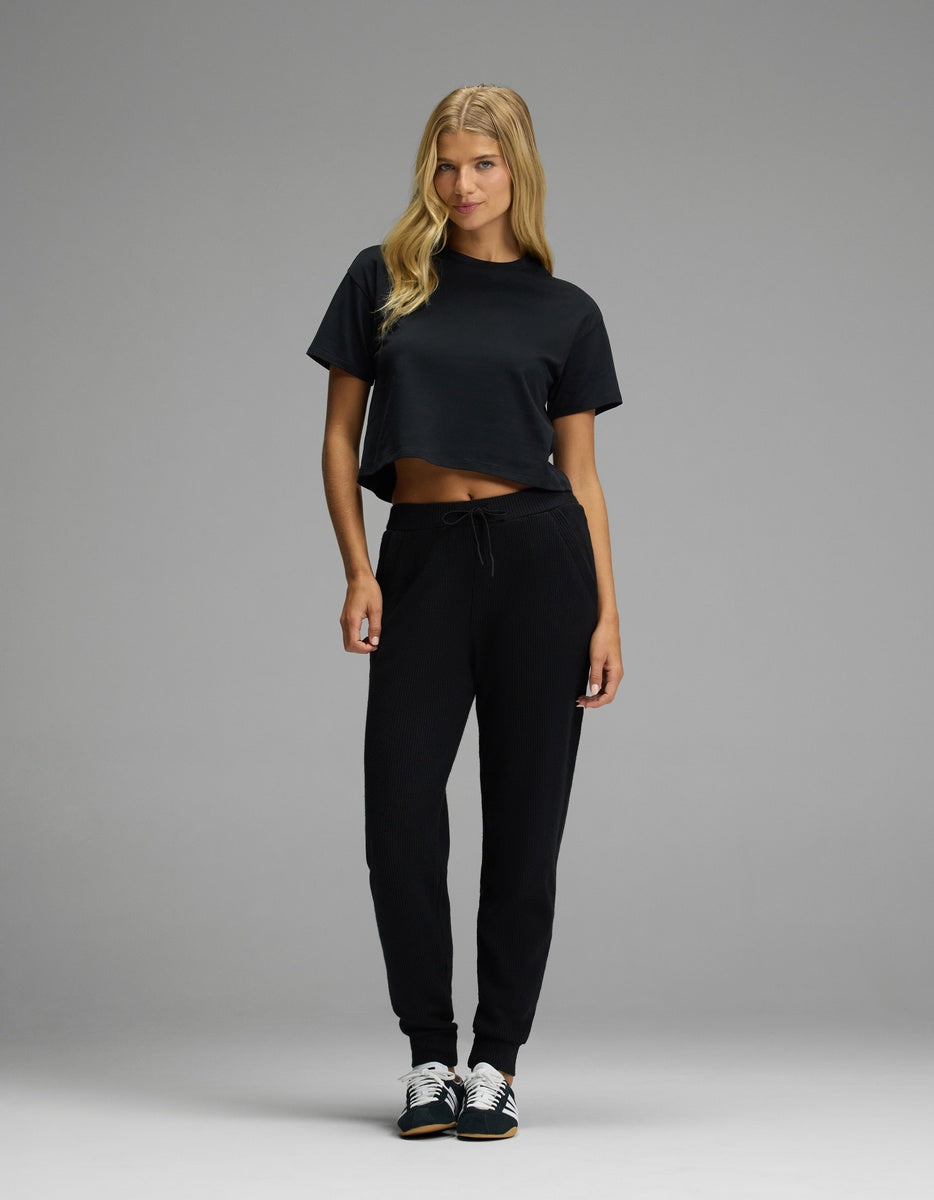A woman wears a black cropped t-shirt and black Cozy Earth Women’s Blake Waffle Joggers with a drawstring waist and ribbed texture, paired with black and white sneakers.|Color:Jet Black