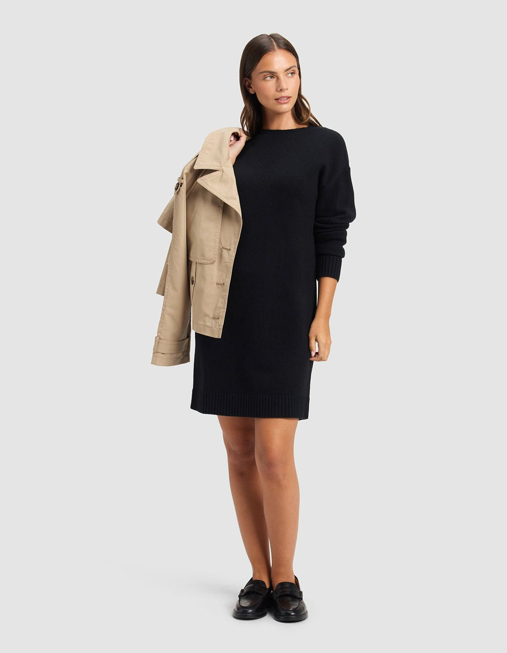 A woman stands against a plain light gray background, wearing Cozy Earth's Women's Brooklyn Sweater Dress in black with black loafers and holding a beige trench coat over her shoulder.|Color:Jet Black