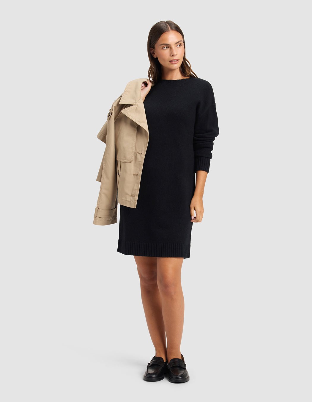 A woman stands against a plain light gray background, wearing Cozy Earth's Women's Brooklyn Sweater Dress in black with black loafers and holding a beige trench coat over her shoulder.|Color:Jet Black