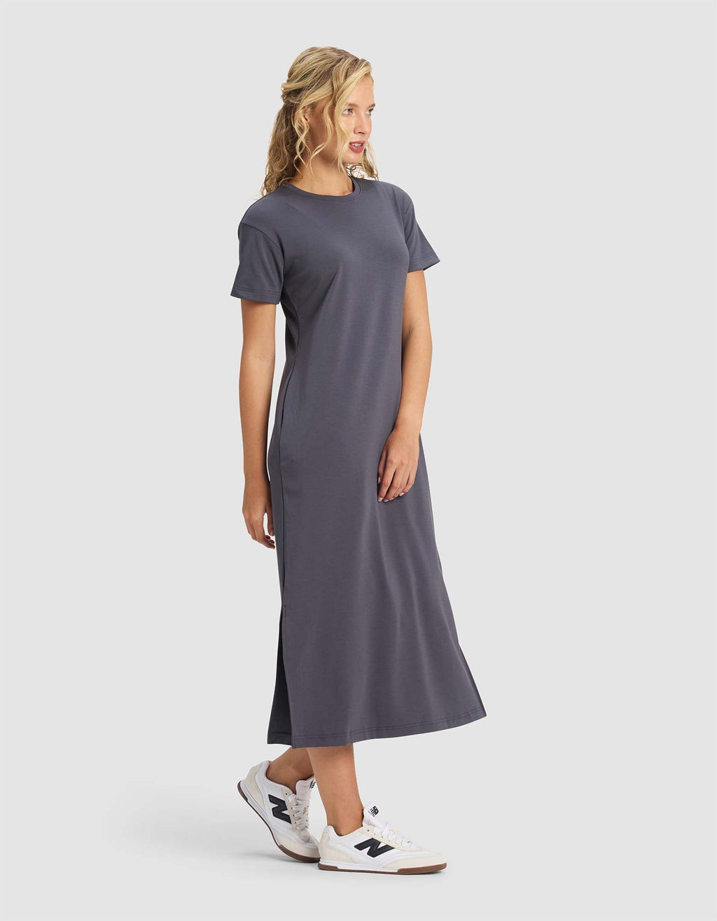A blonde woman stands and smiles, slightly turned to the side, wearing Cozy Earth's Women's Brushed Bamboo Midi Dress in dark gray with short sleeves and white sneakers against a plain light background.|Color:Graphite