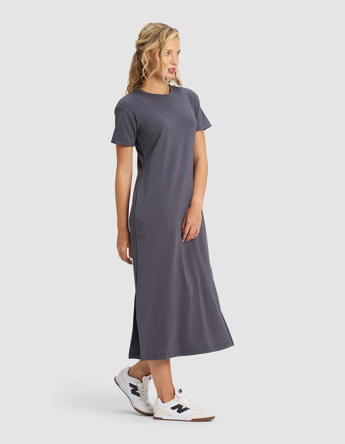 A blonde woman stands and smiles, slightly turned to the side, wearing Cozy Earth's Women's Brushed Bamboo Midi Dress in dark gray with short sleeves and white sneakers against a plain light background.|Color:Graphite