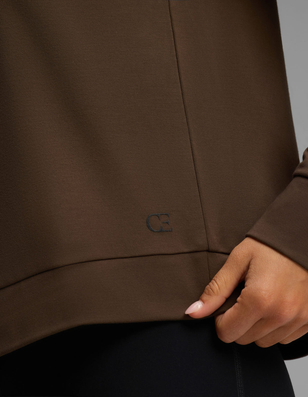 A person wearing the Cozy Earth Women's Brushed Bamboo Pullover Crew in brown gently pinches the hem with one hand, revealing a small, subtle embroidered "CB" logo near the bottom edge.|Color:Espresso