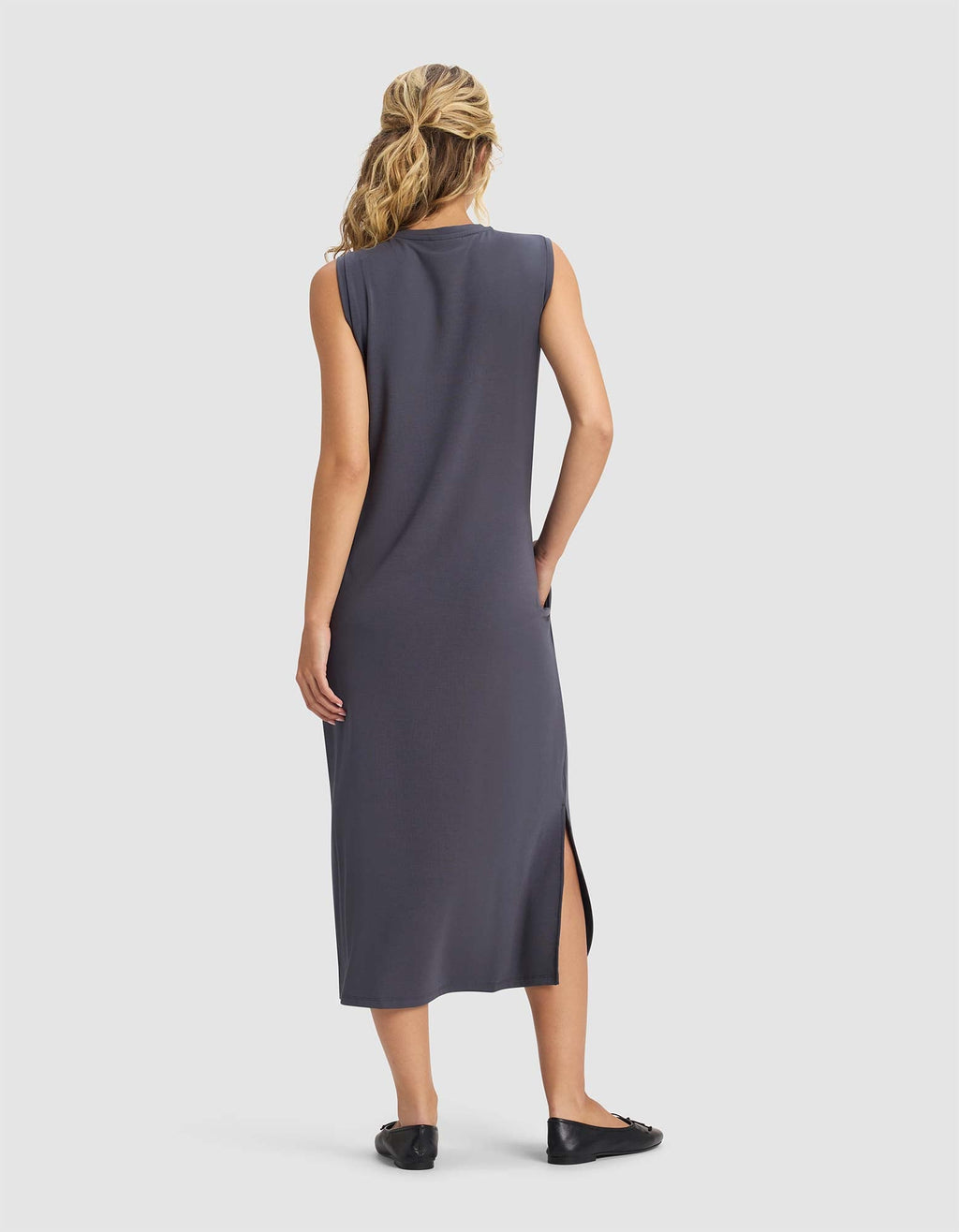 A woman with blonde hair in a ponytail faces away, wearing Cozy Earth's Women's Brushed Bamboo Sleeveless Midi Dress in dark gray with a side slit and black flats, against a plain white background.|Color:Graphite