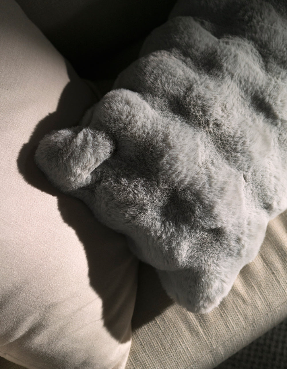 A Cozy Earth Bubble Cuddle Pillow rests on a beige fabric couch, its plush texture highlighted by gentle sunlight that casts soft shadows across the inviting scene.|Color:Light Grey |Size:Body