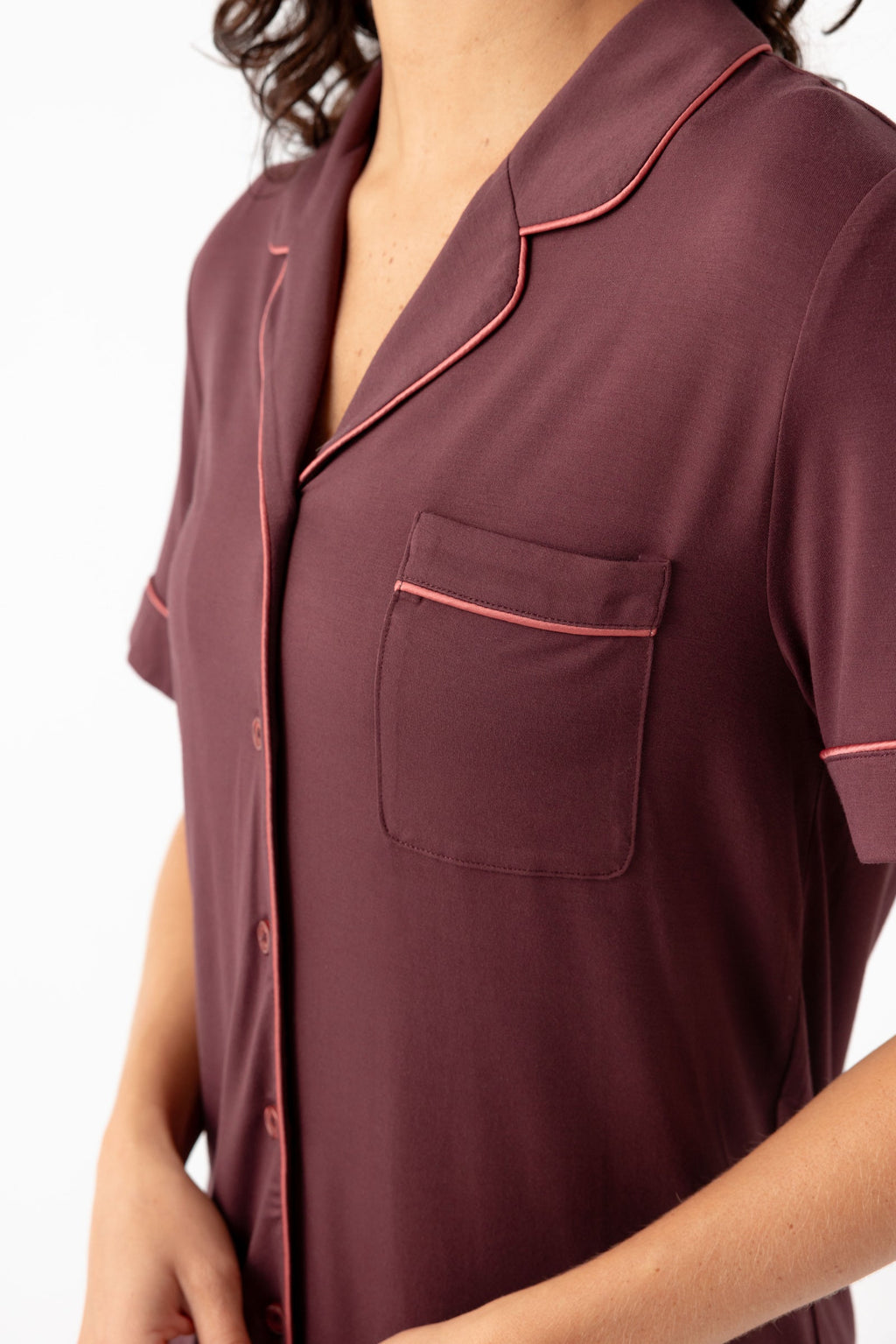 A person wears the Cozy Earth Women's Bamboo Stretch-Knit Short Sleeve Pajama Set—a maroon, pink-piped, button-down top with a breast pocket. The person's head is partially out of frame.|Color:Burgundy