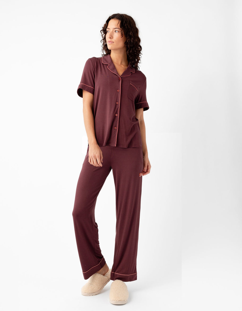 A woman in Cozy Earth's Women's Bamboo Stretch-Knit Short Sleeve & Pant Pajama Set, featuring a collar, buttons, and loose-fitting pants in burgundy, stands against a white background wearing beige slippers and looking to the side with one hand on her hip.|Color:Burgundy