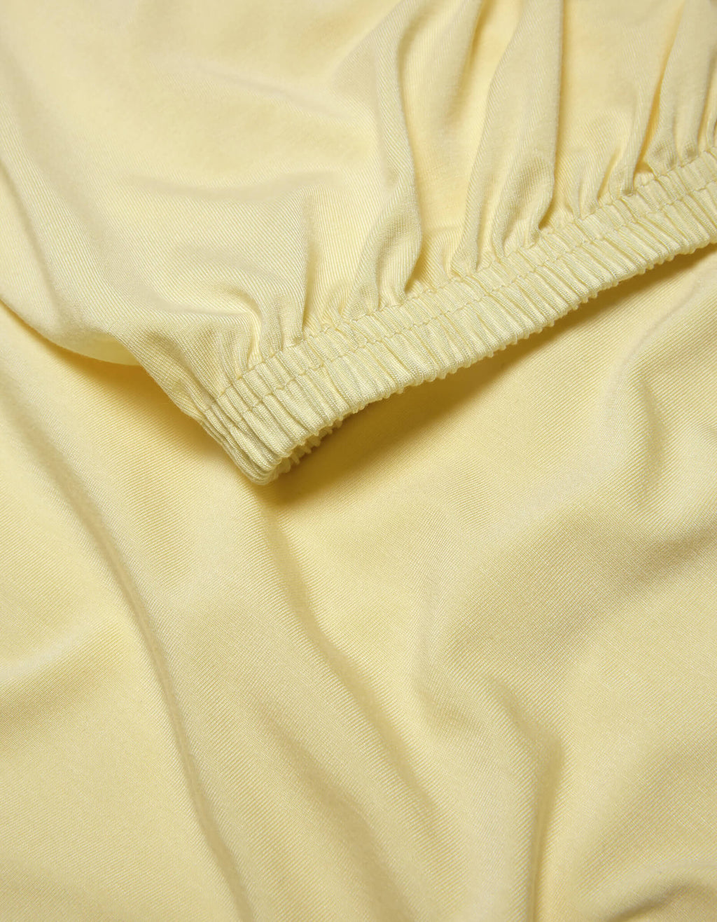 A close-up of the Cozy Earth Bamboo Jersey Fitted Sheet shows light yellow fabric with elasticized, gathered edges that create gentle folds and a smooth, soft texture.|Color:Meringue