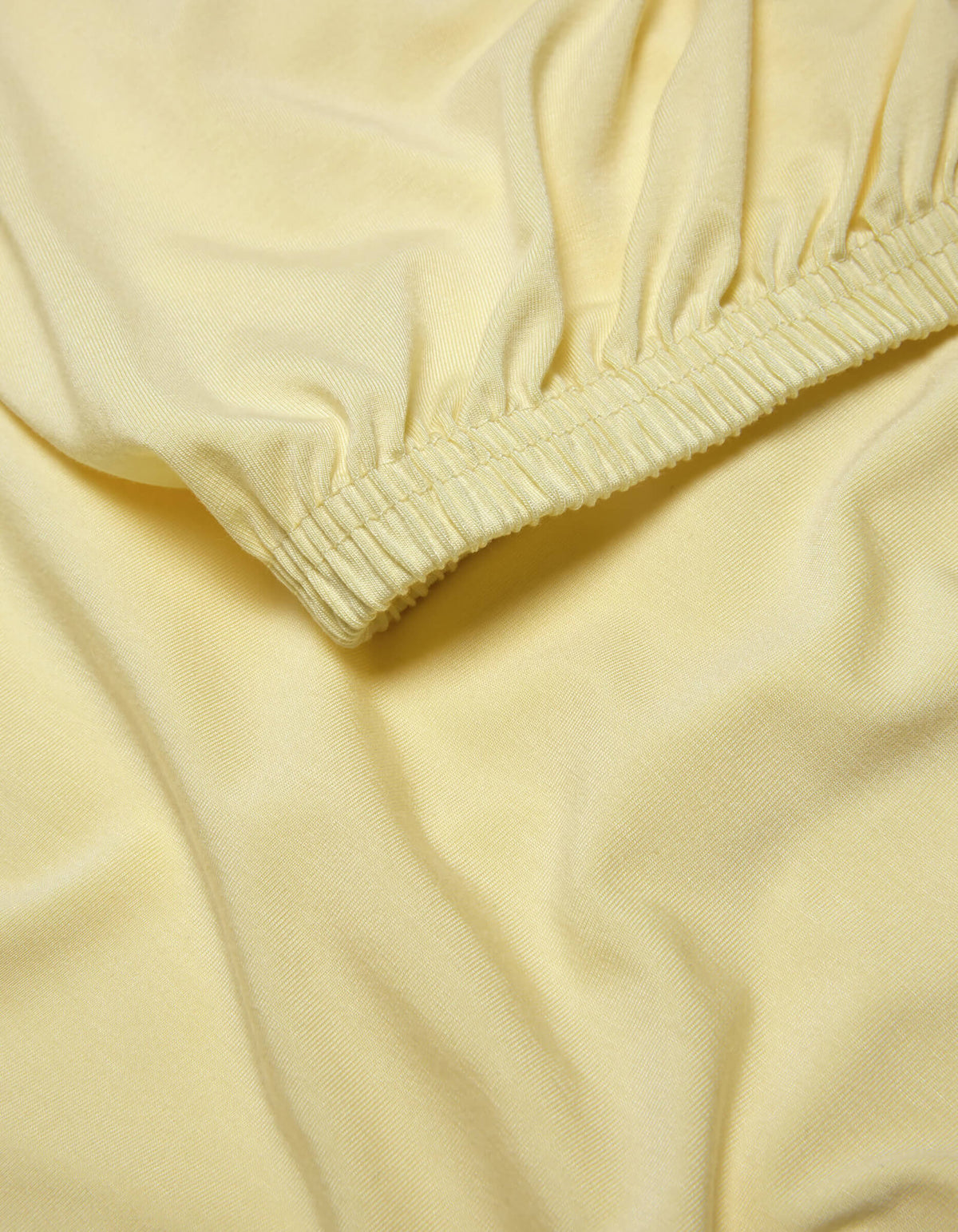 A close-up of the Cozy Earth Bamboo Jersey Fitted Sheet shows light yellow fabric with elasticized, gathered edges that create gentle folds and a smooth, soft texture.|Color:Meringue