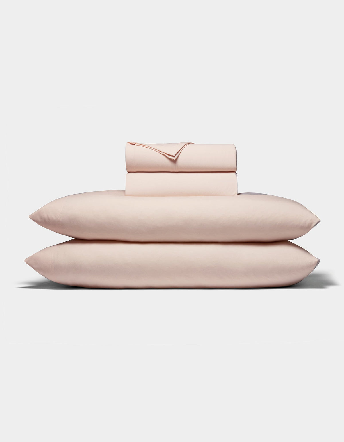 A neatly folded stack of Cozy Earth's Bamboo Jersey Sheet Set, in pale pink, featuring two pillows, a fitted sheet, a flat sheet, and pillowcases arranged on a white background.|Color:Petal
