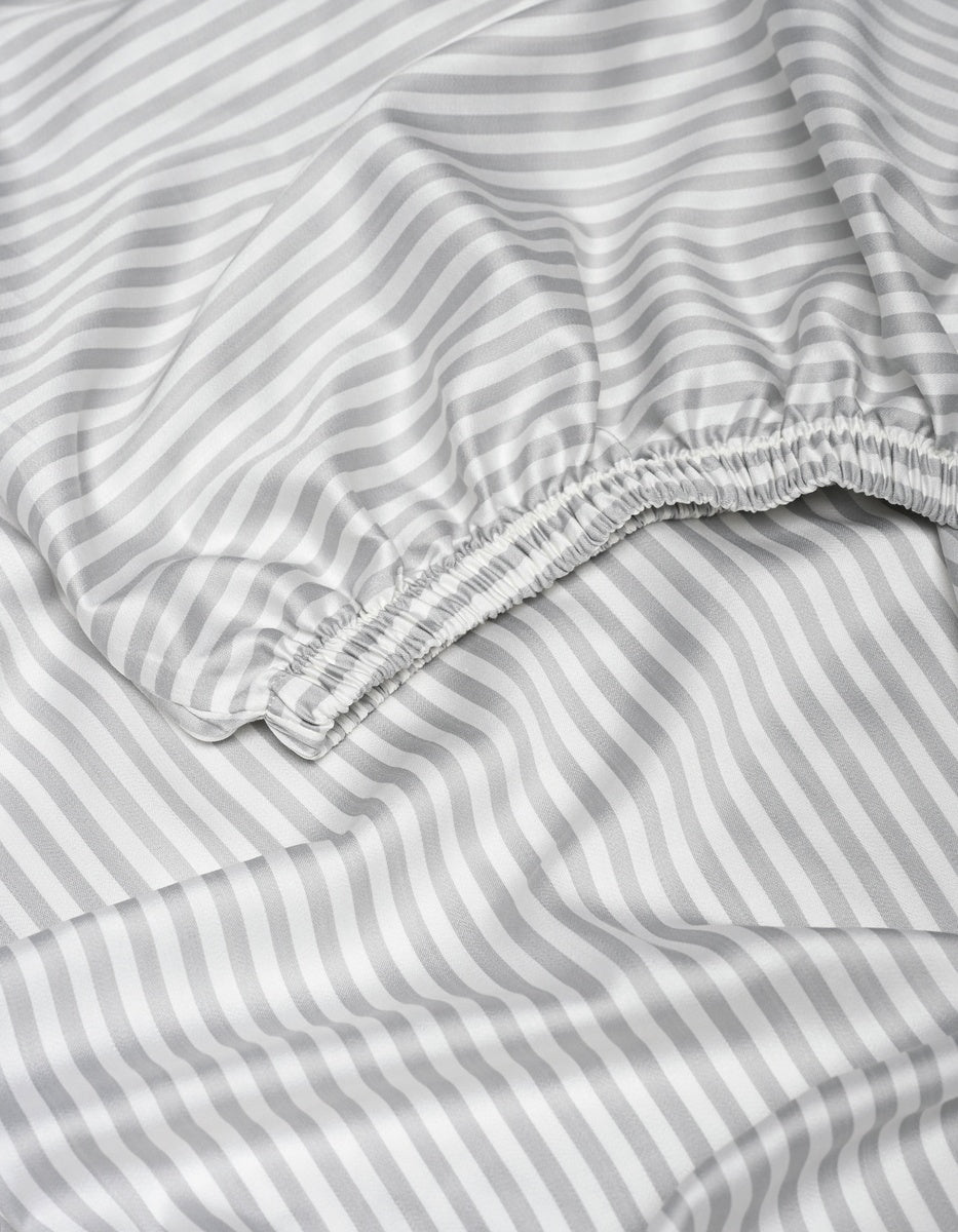 A close-up of the Cozy Earth Bamboo Fitted Sheet displays its gray and white diagonal stripes, featuring a gathered elastic edge and smooth, slightly wrinkled fabric.|Color:Classic Stripe in Dew