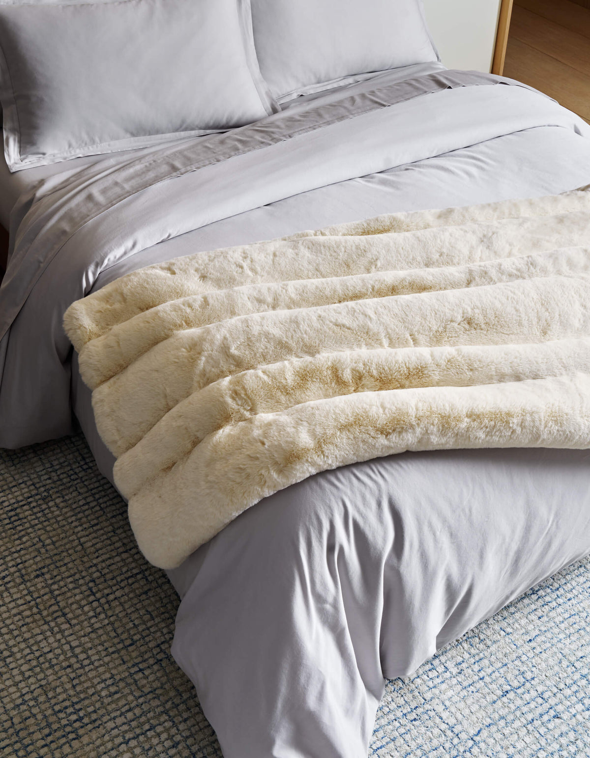 A neatly made bed with light gray sheets and pillowcases is topped with Cozy Earth’s Channel Cuddle Blanket in cream, draped at the foot. A blue and white textured rug is partially visible on the floor.|Color:Creme