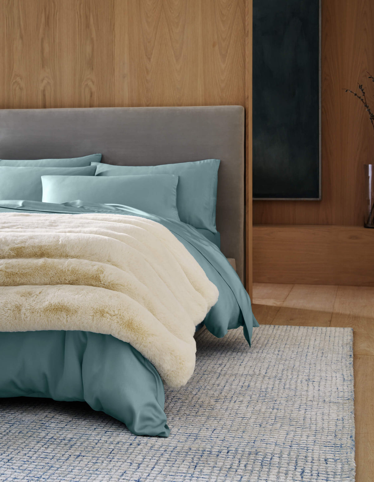A modern bedroom with a light gray upholstered bed, teal bedding, and the Cozy Earth Channel Cuddle Blanket in cream. Wooden walls, a light blue textured rug, minimal decor, and a tall vase with branches complete the look.|Color:Creme