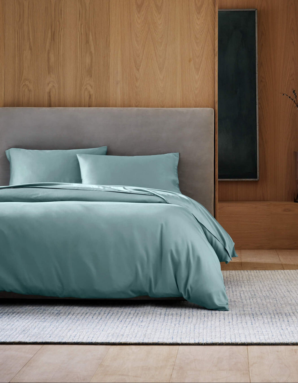 A modern bedroom showcases the Cozy Earth Bamboo Bedding Deluxe Bundle in light teal, paired with a gray upholstered headboard, a light rug, and minimalist wood-paneled walls.|Color:River