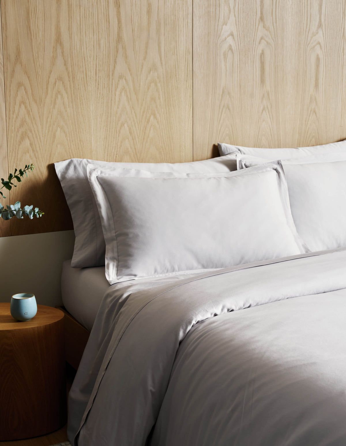 A bed featuring the Cozy Earth Soft-Wash Cotton Duvet Cover in light gray is set against a wood-paneled wall. A small round wooden nightstand displays a blue candle and greenery arranged in a white vase.|Color:Dew