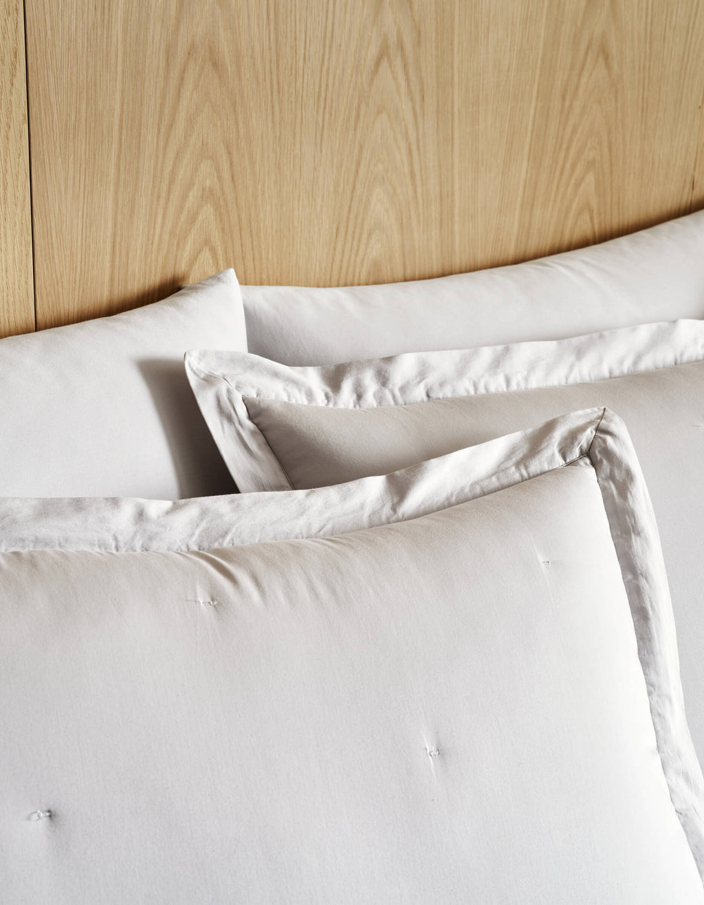 Three large Soft-Wash Cotton Quilted Shams by Cozy Earth, in white with subtle tufting, are stacked against a light wood headboard for a cozy, minimalist bed setting.|Color:Dew