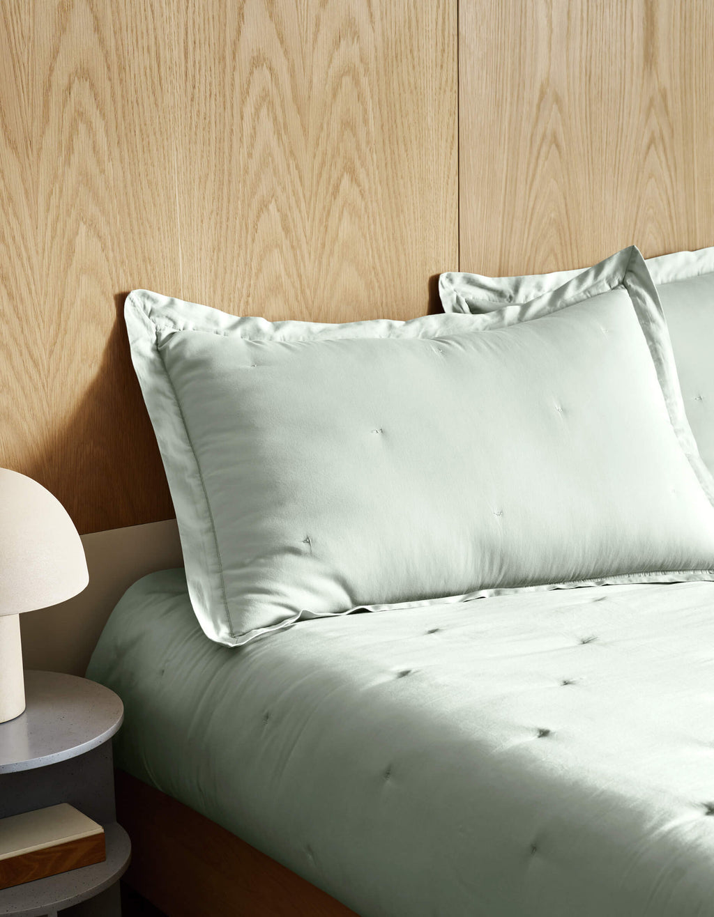 A neatly made bed features Cozy Earth's Soft-Wash Cotton Quilt in light sage green, set against a wooden paneled wall. On the left, a round bedside table holds a modern white lamp.|Color:Laurel