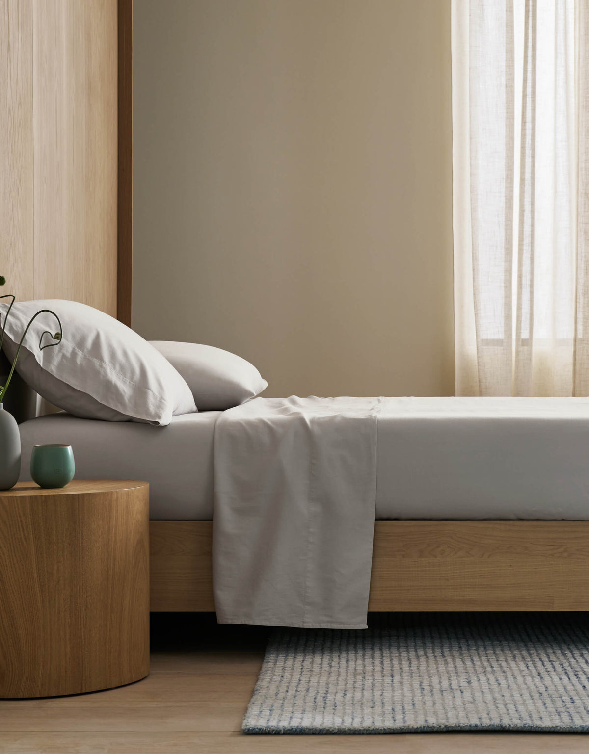 A minimalist bedroom features a neatly made bed with Cozy Earth's Soft-Wash Cotton Flat Sheet, light gray bedding, a wooden nightstand with a green vase and cup, a light blue-gray rug, and sheer curtains letting in gentle natural light.|Color:Dew