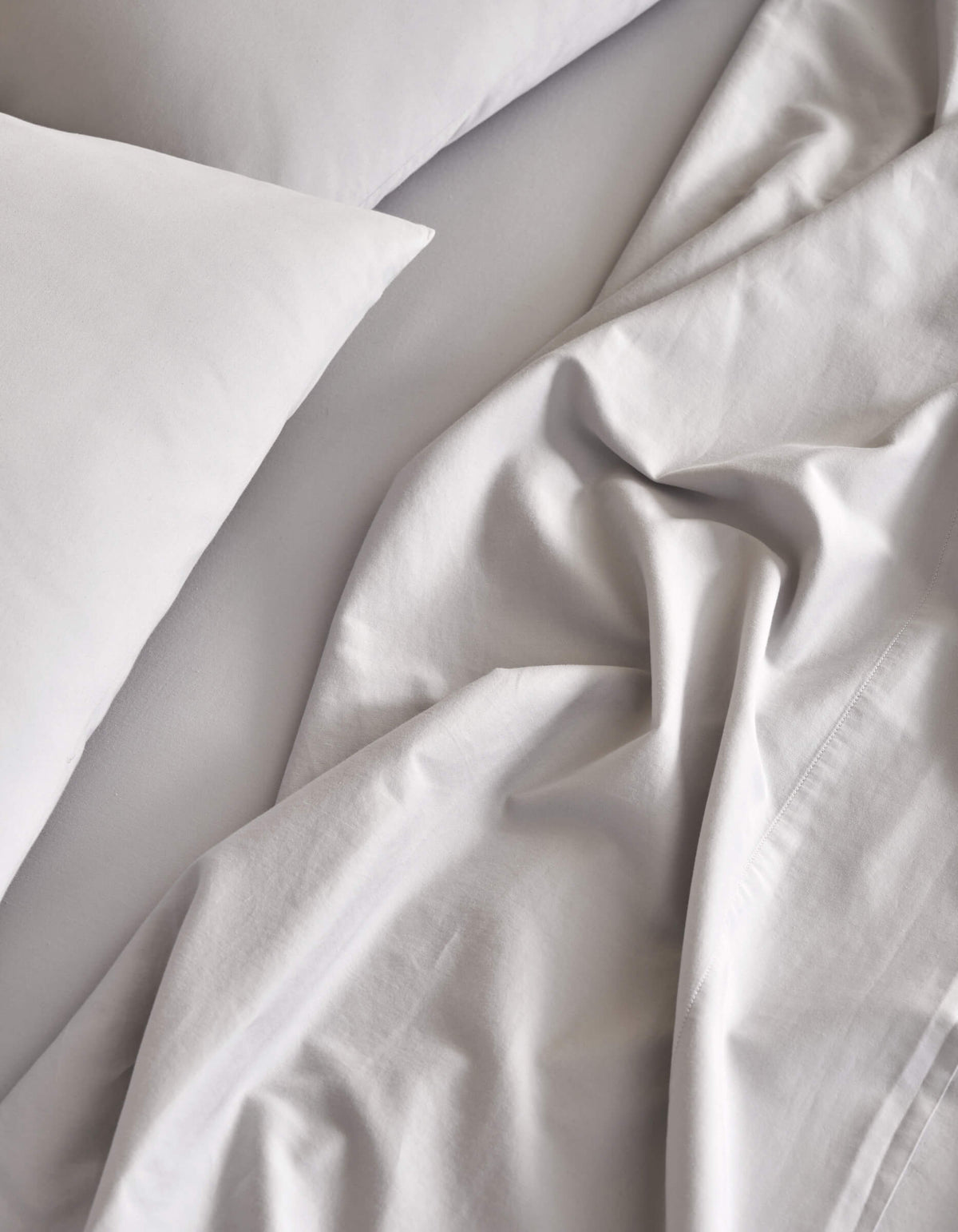 A close-up view of a bed featuring Cozy Earth's Soft-Wash Cotton Sheet Set in light gray. The soft, slightly wrinkled fabric creates a relaxed and inviting look.|Color:Dew