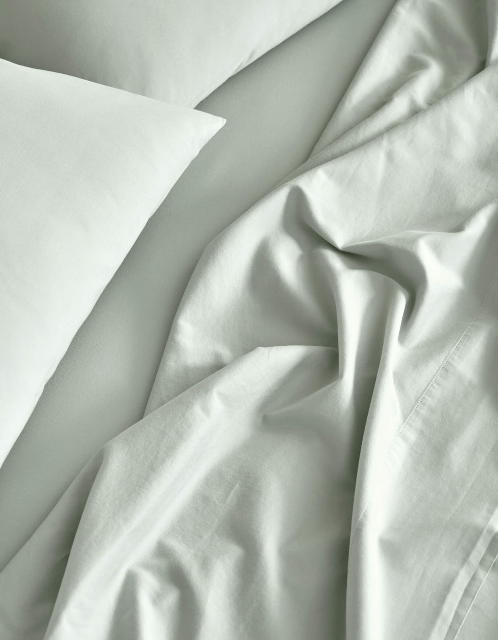 A close-up of a neatly made bed features the Cozy Earth Soft-Wash Cotton Sheet Set in light gray, with slightly rumpled sheets and pillowcases creating soft folds and natural shadows.|Color:Laurel