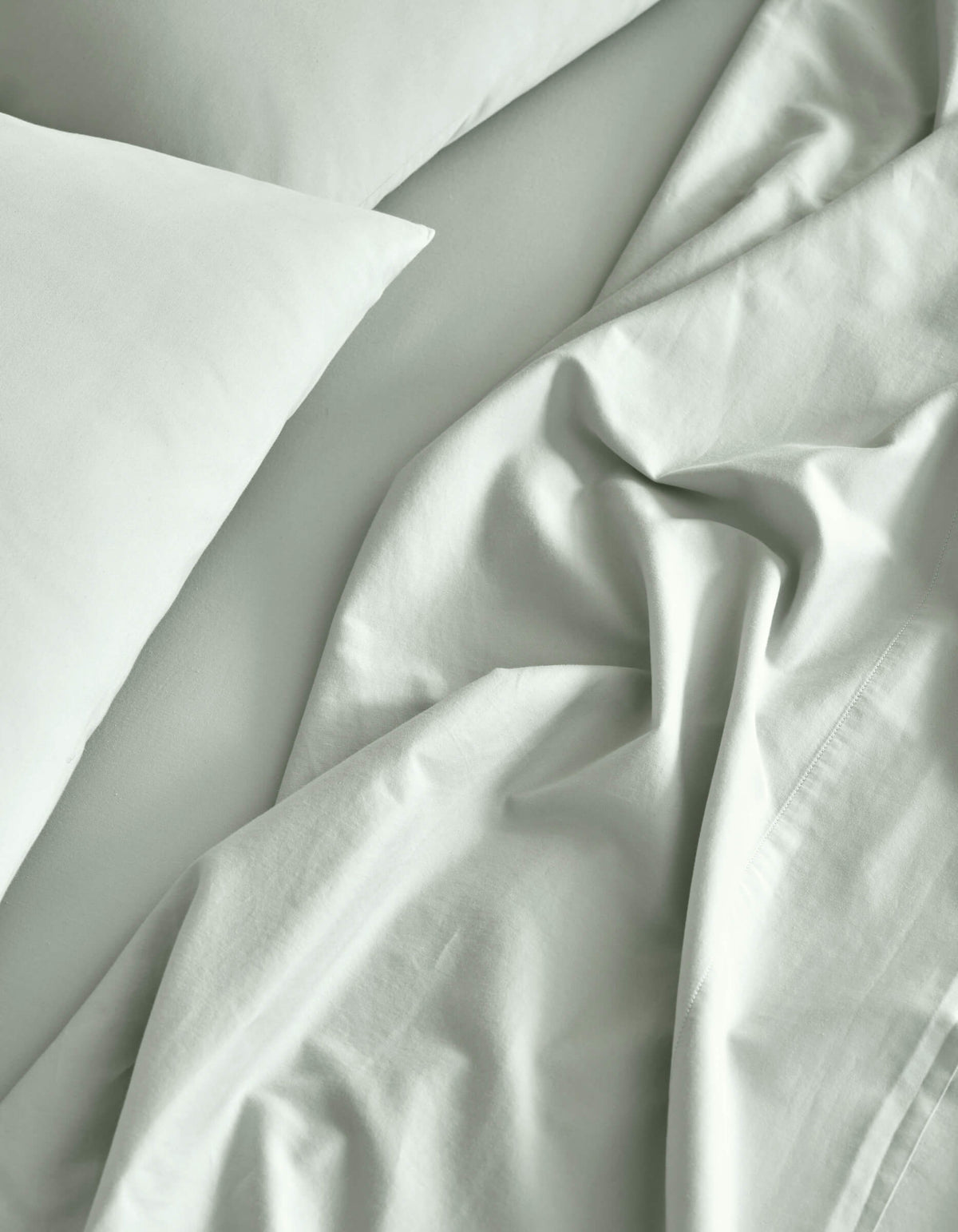 A close-up of a neatly made bed features the Cozy Earth Soft-Wash Cotton Sheet Set in light gray, with slightly rumpled sheets and pillowcases creating soft folds and natural shadows.|Color:Laurel