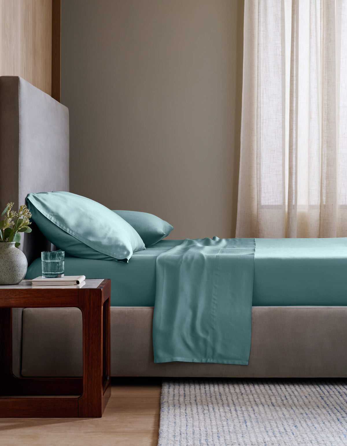 A neatly made bed with Cozy Earth's Bamboo Fitted Sheet in light teal and matching pillows sits beside a wooden nightstand holding a glass of water and flowers, all in a softly lit, modern bedroom with beige walls and curtains.|Color:River