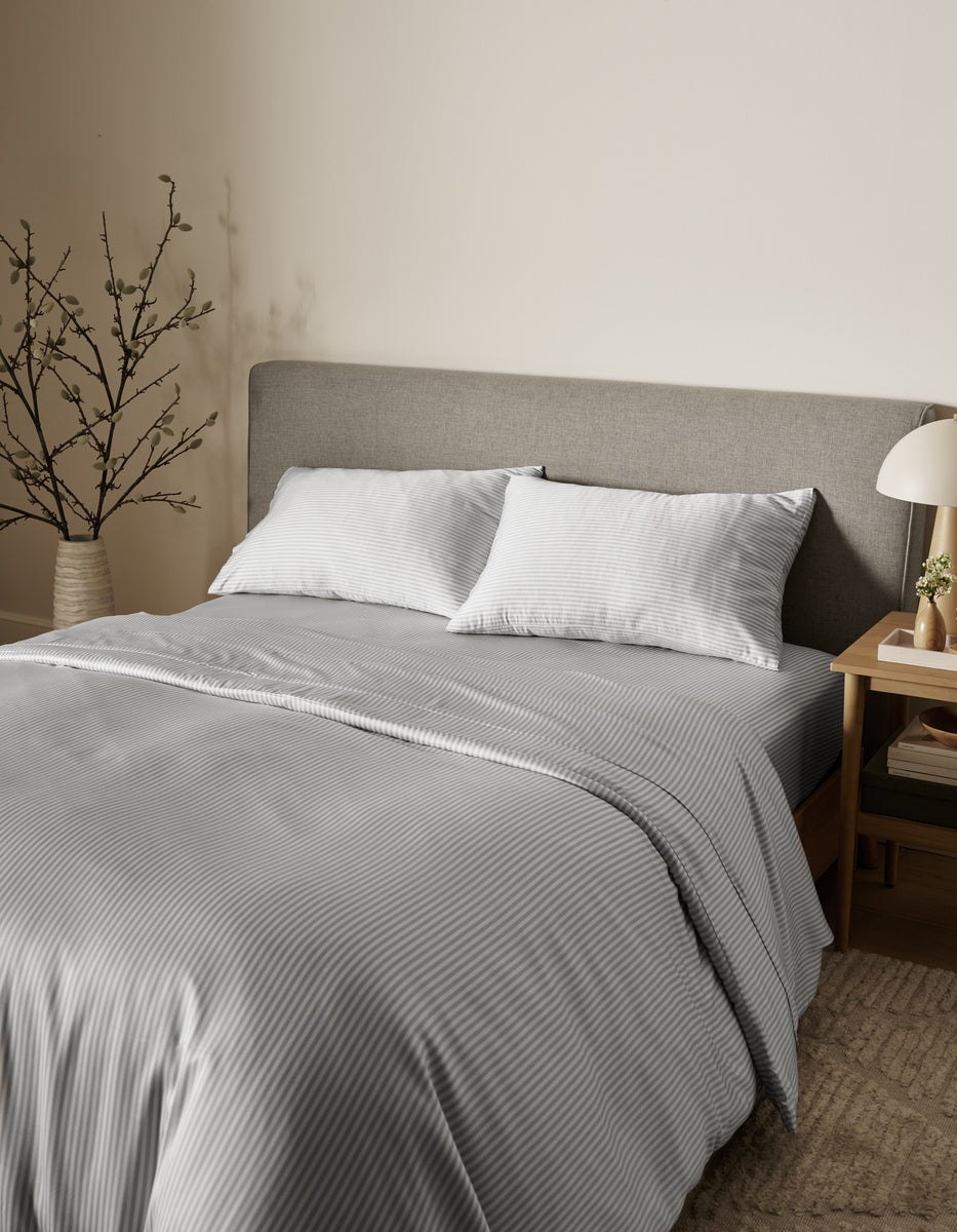 A neatly made bed featuring the Cozy Earth Bamboo Duvet Cover in gray and white stripes, styled with two pillows, a gray upholstered headboard, a small bedside table with a lamp, and a vase of branches in a cozy, neutral-toned bedroom.|Color:Classic Stripe in Dew