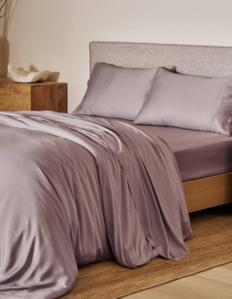 A bed dressed in a silky mauve-lavender bedding set with matching pillowcases, styled in a warm bedroom setting with a wooden nightstand and textured headboard. |Color:Lavender