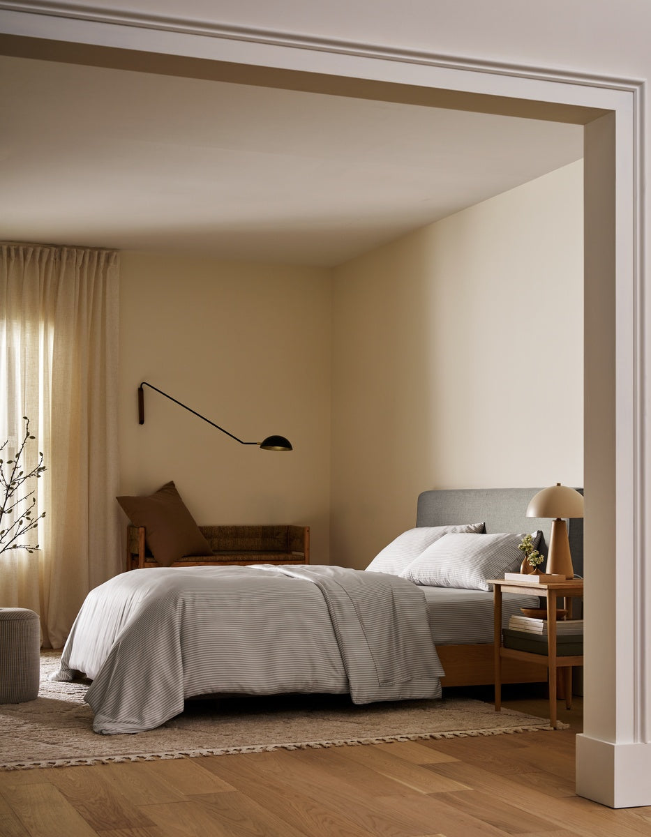 A minimalist bedroom interior with a neatly made bed dressed in white linens, a gray upholstered headboard, warm beige walls, wood flooring, a bedside table with a lamp, and a wall-mounted black lamp.|Color:Classic Stripe in Dew