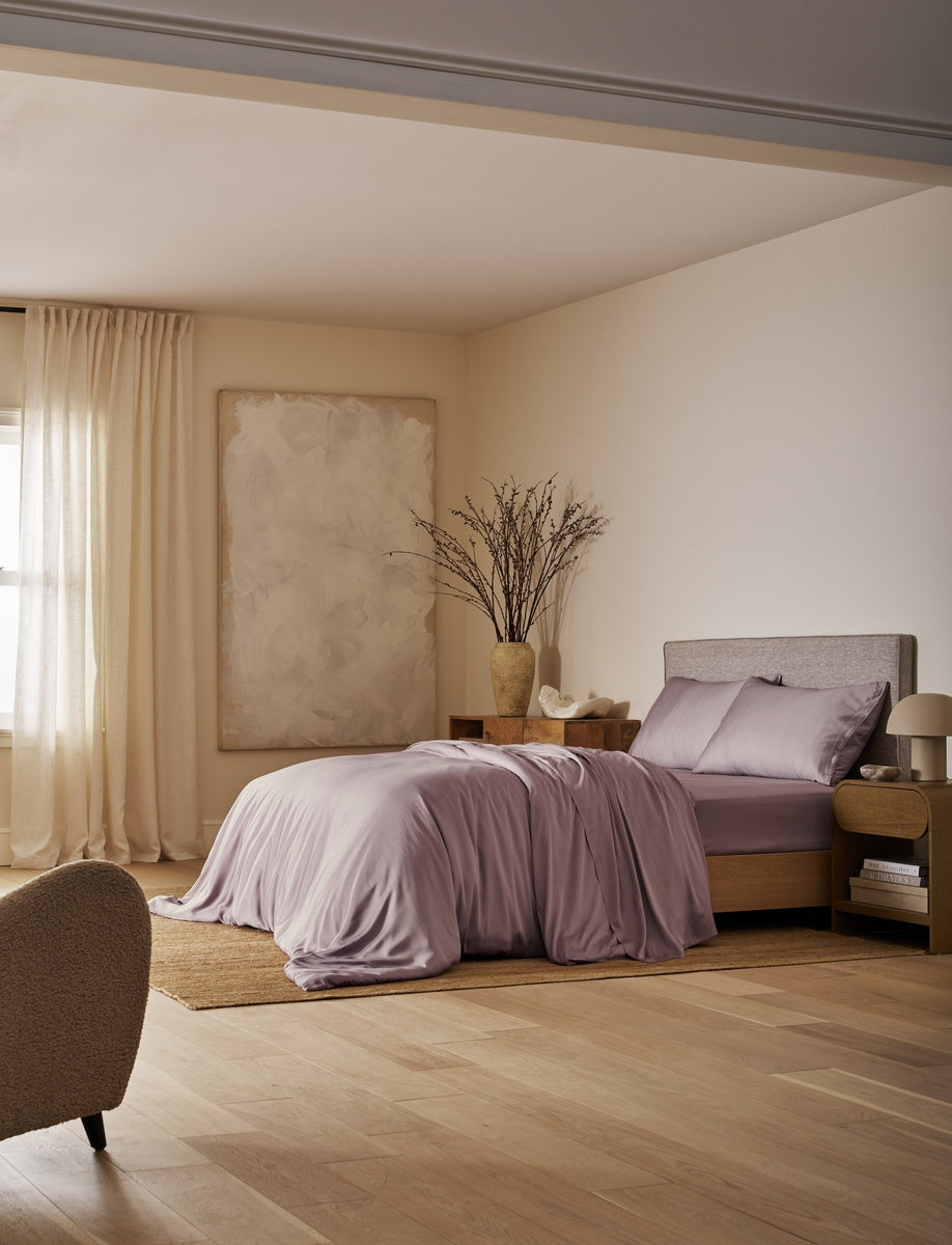 A bedroom scene with a bed dressed in soft lavender bedding, set in a warm neutral room with beige walls, wood flooring, curtains, and a large abstract painting.|Color:Lavender