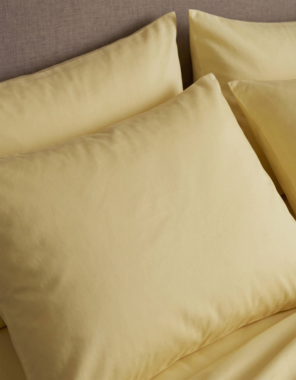 Close-up of neatly arranged Cozy Earth Bamboo Jersey Pillowcases in pale yellow on a bed with a beige upholstered headboard.|Color:Meringue