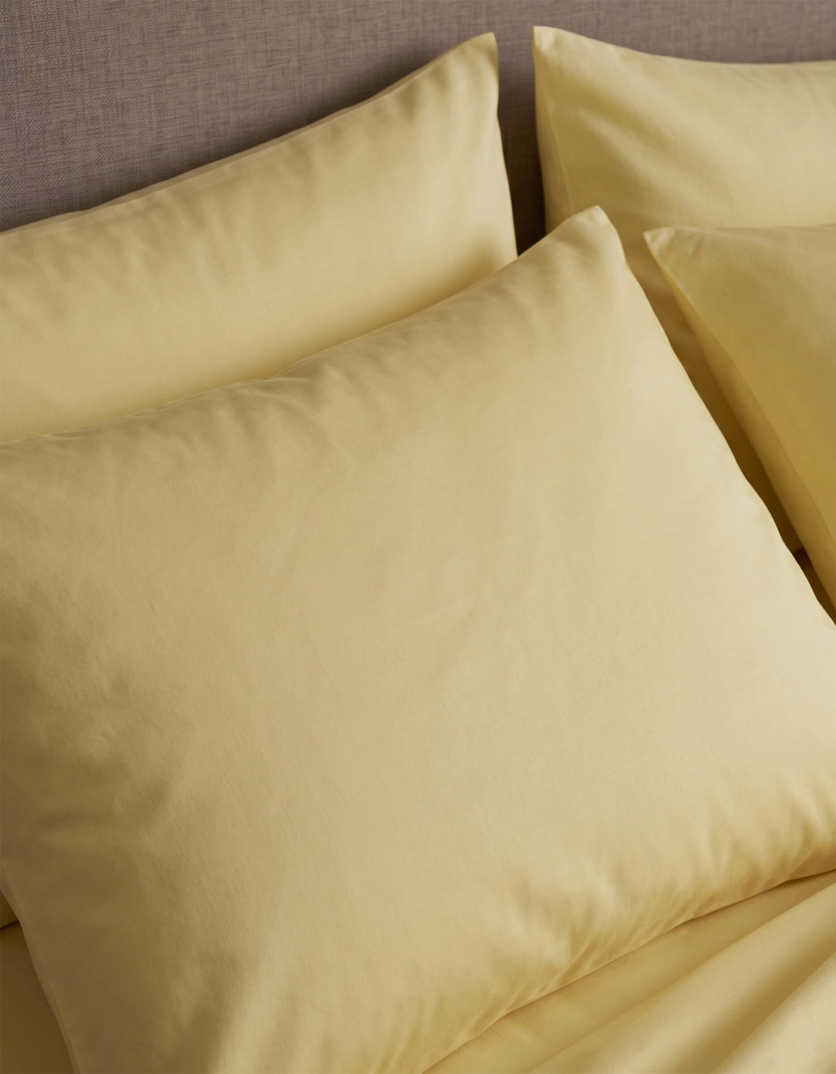 Close-up of neatly arranged Cozy Earth Bamboo Jersey Pillowcases in pale yellow on a bed with a beige upholstered headboard.|Color:Meringue
