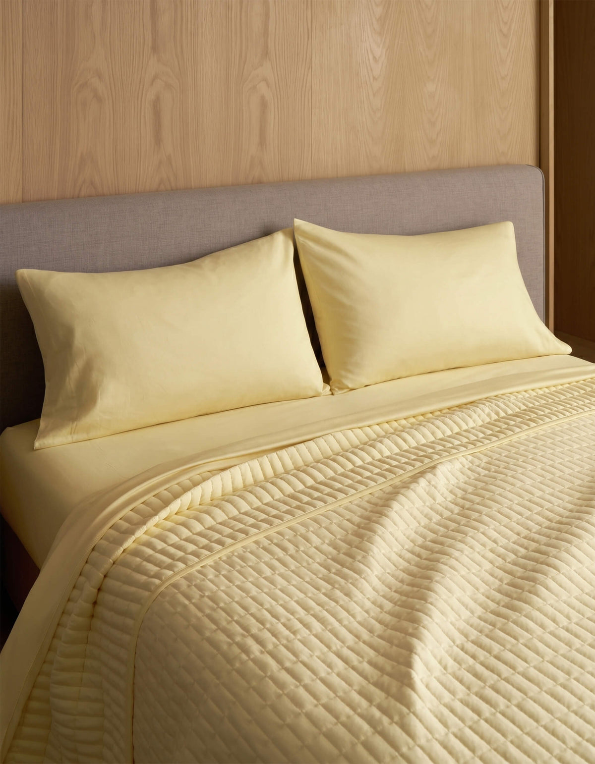 A neatly made bed features Cozy Earth Bamboo Jersey Pillowcases, two matching pillows, and a quilted yellow blanket, all set against a wood-paneled wall and a light gray upholstered headboard.|Color:Meringue