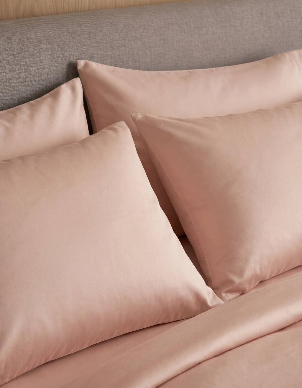 A close-up of a neatly made bed with Cozy Earth Bamboo Jersey Pillowcases in light pink, paired with matching sheets and set against a grey upholstered headboard.|Color:Petal