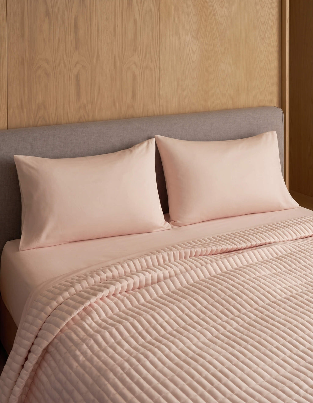 A neatly made bed features two Cozy Earth Bamboo Jersey Pillowcases in light pink, matching sheets, a quilted pink blanket, and a gray upholstered headboard set against a wooden wall.|Color:Petal